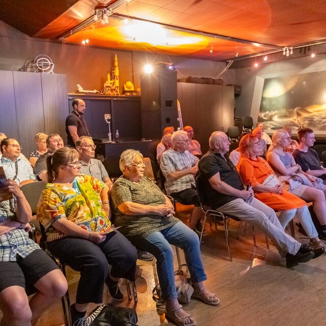 Besucherinnen und Besucher hören einen Vortrag im abgedunkelten Saal.